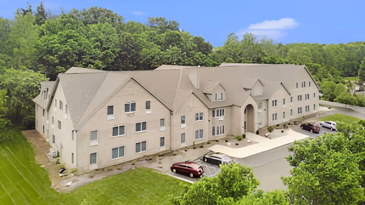 Three-story beige senior living building with an arched main entrance, parking area with cars, and trees surrounding the property.