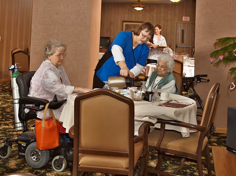 A staff member pours coffee for two elderly women seated at a dining table in a senior living facility dining room.