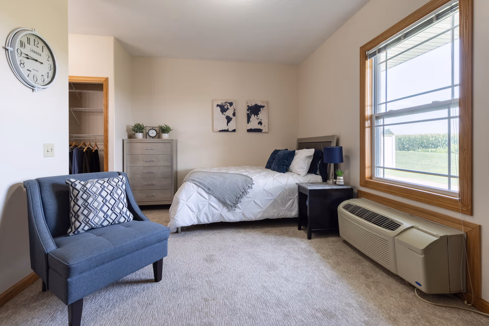A bright furnished bedroom with a bed, armchair, dresser, nightstand, and large window overlooking a lawn.