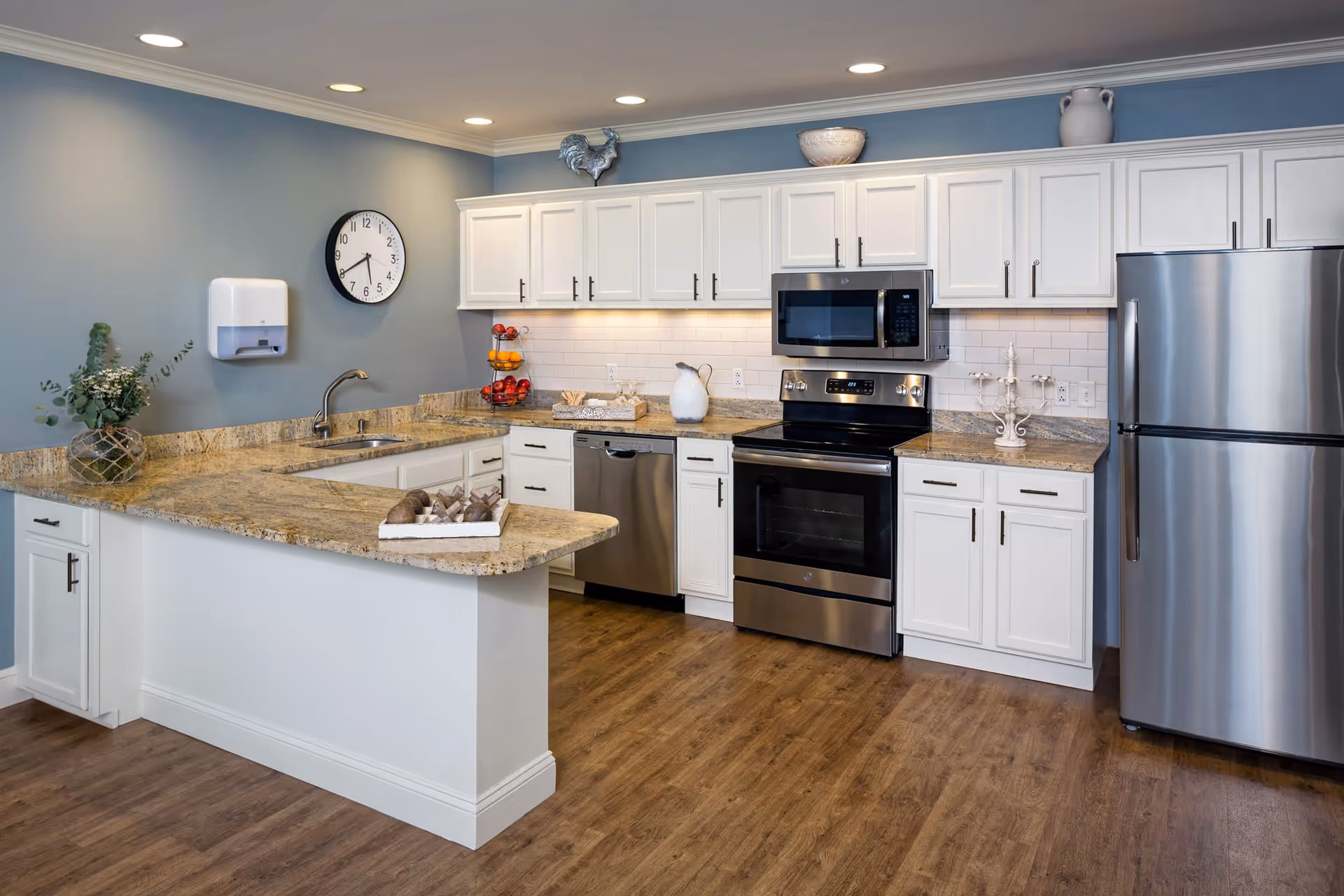 A modern kitchen with white cabinets, granite countertops, and stainless steel appliances including a refrigerator, oven, microwave, and dishwasher. The kitchen has a light blue wall with a clock, a paper towel dispenser, and decorative items such as a vase with greenery and a fruit stand. The floor is wood, and the space is well-lit with recessed ceiling lights.