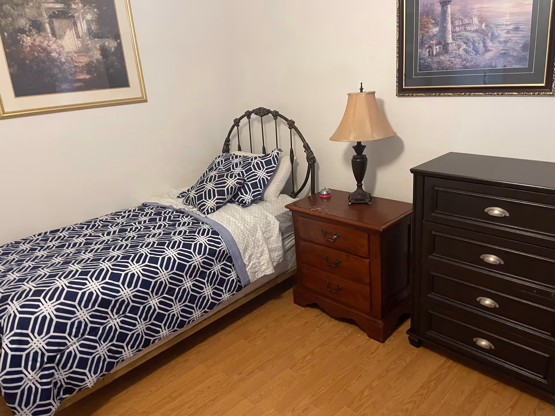 A small bedroom with a single bed featuring a dark metal headboard and blue and white patterned bedding. Next to the bed is a wooden nightstand with a lamp and a small red object on it. There is a dark wooden dresser with silver handles to the right of the nightstand. Two framed paintings hang on the light-colored walls above the bed and dresser. The floor is wooden.