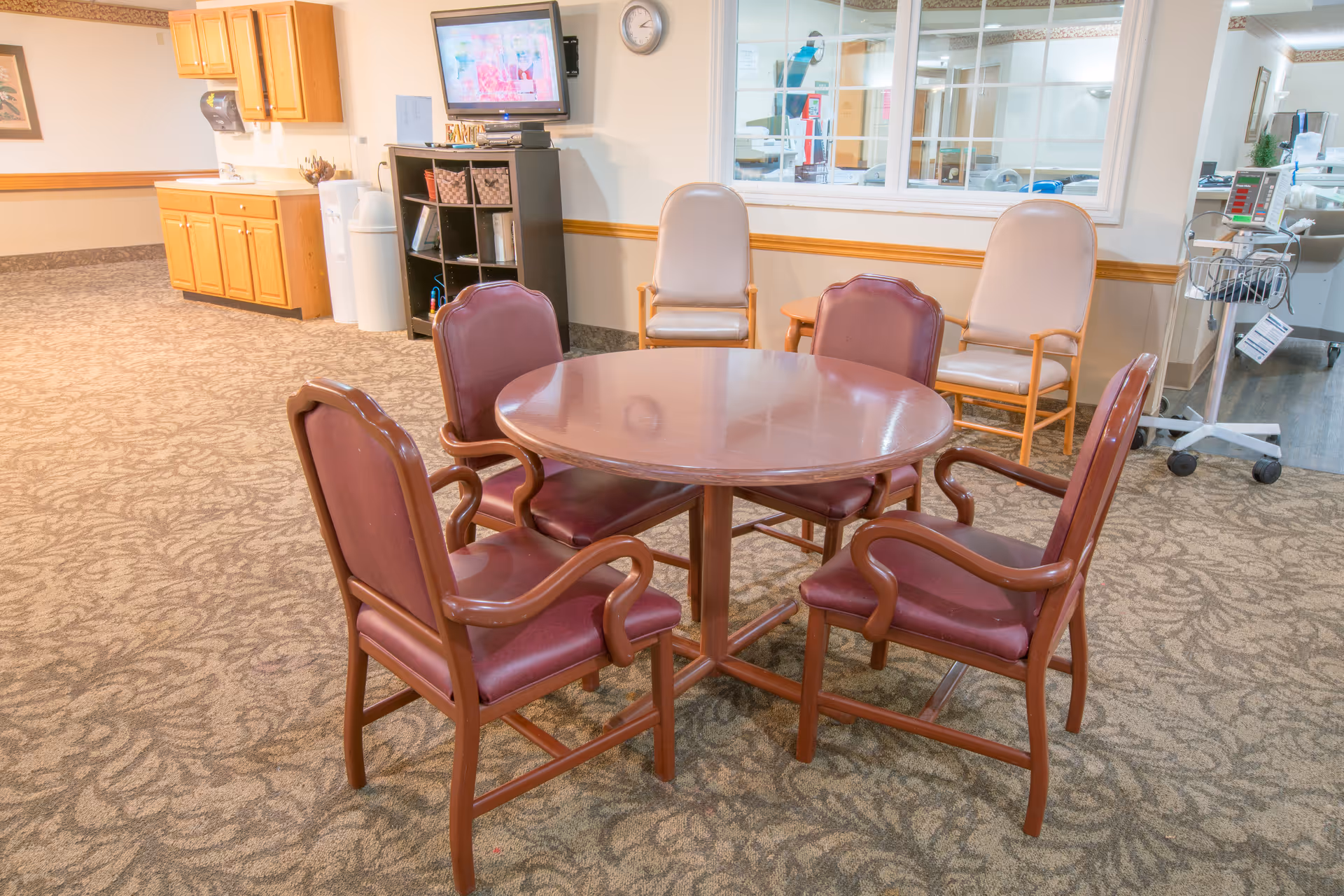 A round wooden table surrounded by four maroon cushioned chairs in a carpeted room. Behind the table are two beige cushioned chairs and a small wooden side table. A TV is mounted on a black shelving unit with baskets and books. Wooden cabinets and a water dispenser are visible on the left side, and a medical area with equipment is visible through a large window on the right.