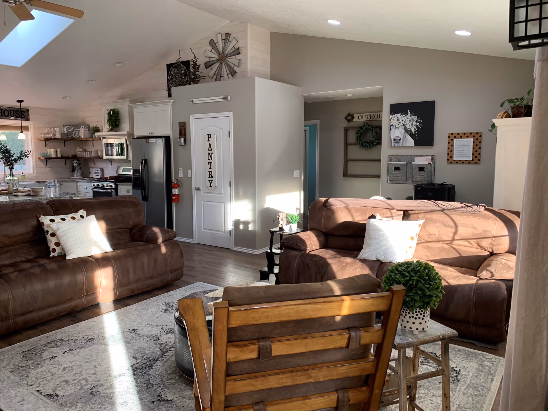 A cozy living room area with two brown leather sofas, a wooden chair, and a small side table with a potted plant. The room has a light-colored rug and is well-lit by natural sunlight. In the background, there is a kitchen with white cabinets, a stainless steel refrigerator, a microwave, and open shelves with decor items. A door labeled 'PANTRY' is visible, along with wall decorations including a windmill and a cow painting.