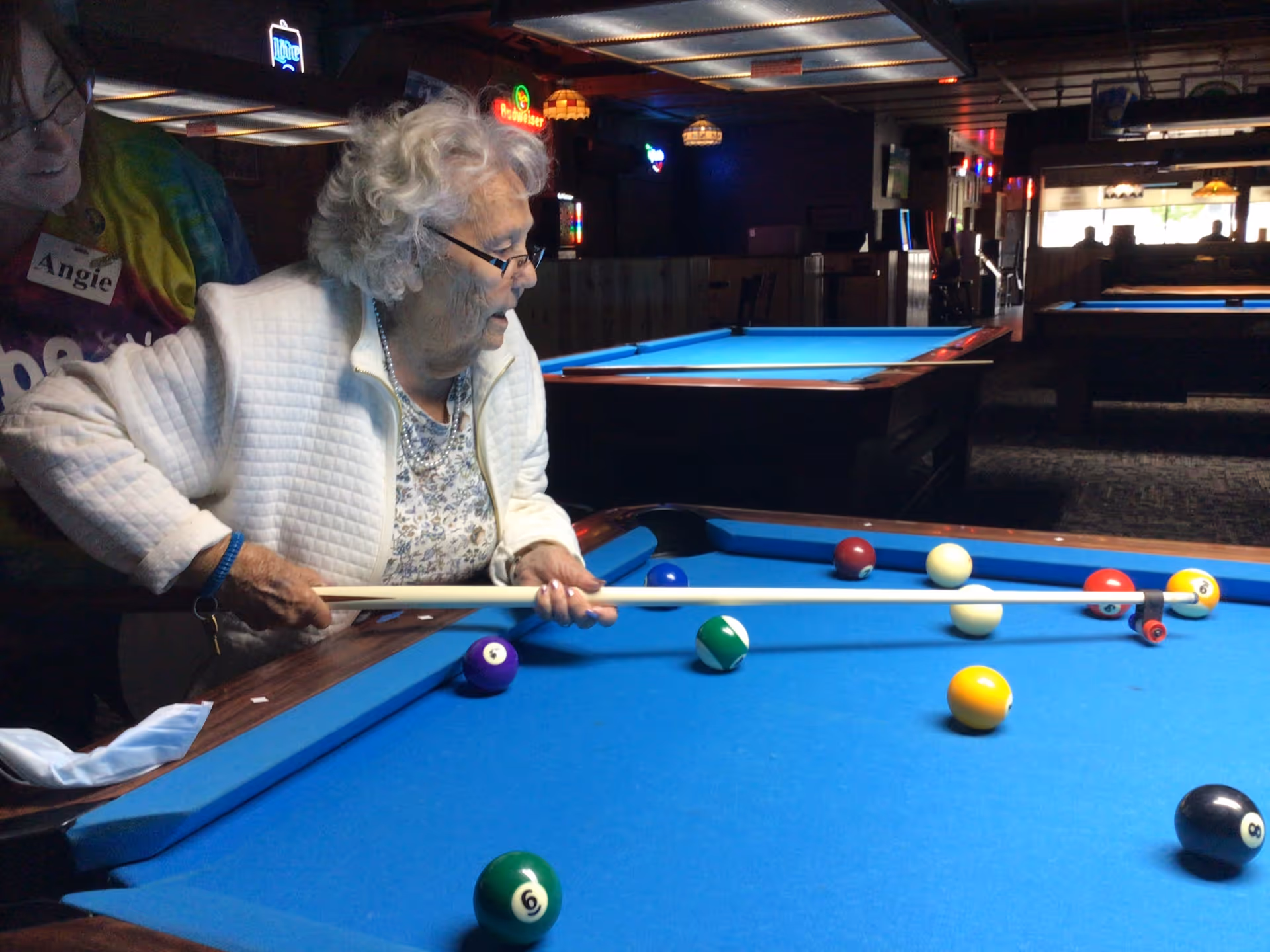 An elderly woman with white hair and glasses is playing pool in a dimly lit billiards room. She is holding a pool cue and aiming at the balls on a blue pool table. Another person wearing a colorful shirt and a name tag that reads 'Angie' is partially visible beside her. Several other pool tables and hanging lights are visible in the background.