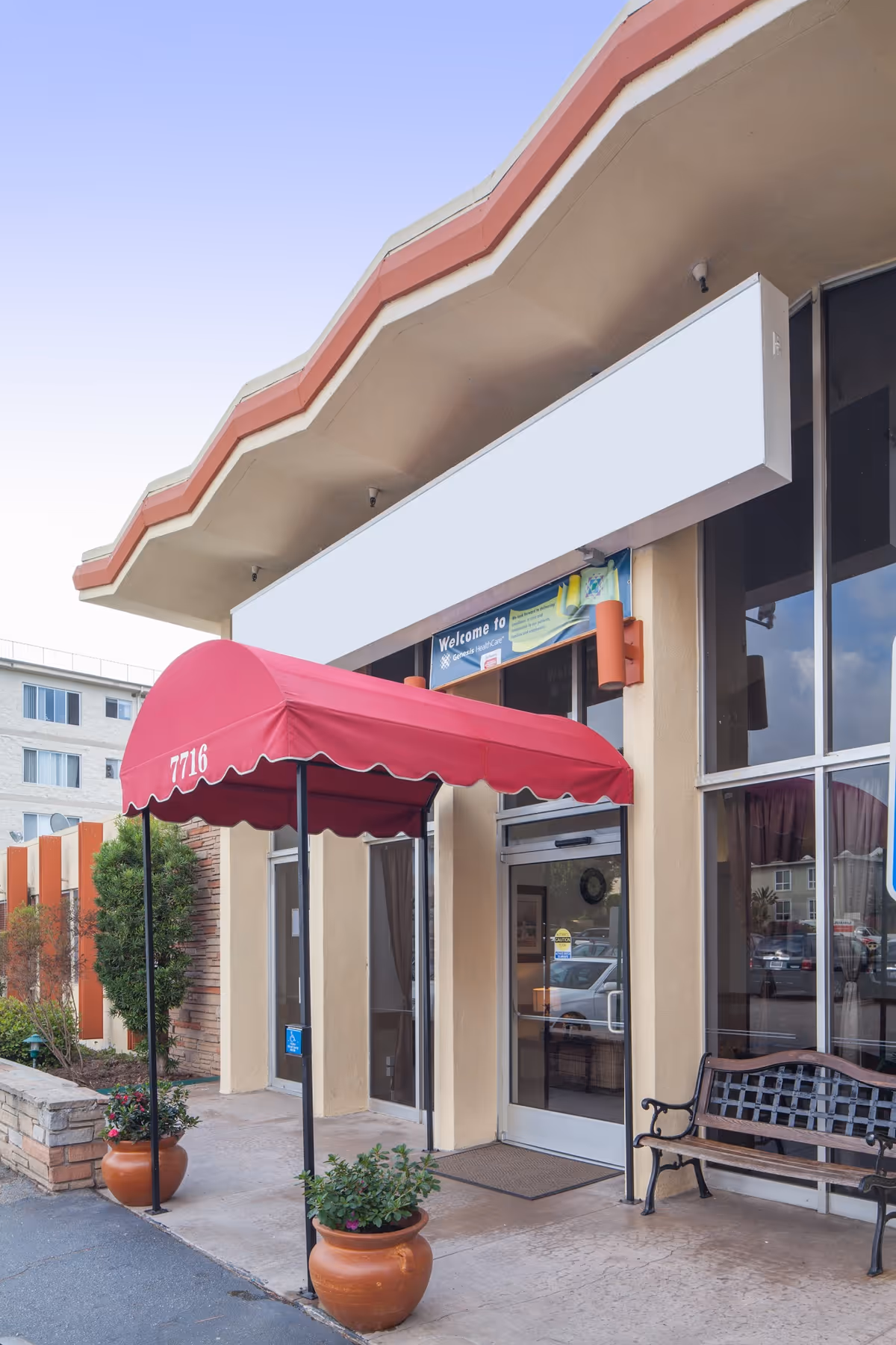 Front entrance of Playa del Rey Center showing a red canopy marked "7716" over glass doors, potted plants, and a bench.