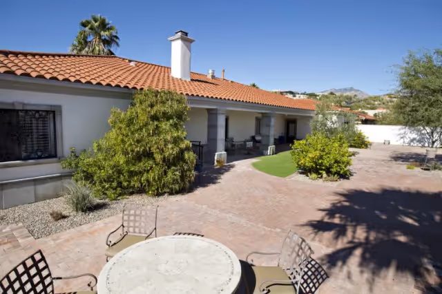 Sunny courtyard with a round patio table and chairs in front of a single-story stucco building with a tiled roof and covered porch.