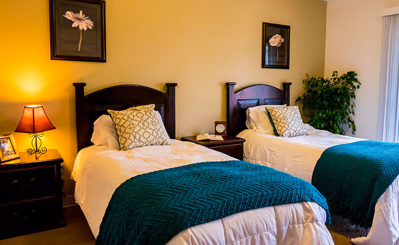 A bedroom with two twin beds featuring dark wooden headboards, white bedding, patterned pillows, and teal blankets. There is a nightstand between the beds with a clock and a phone, a lamp on a side table to the left, framed floral artwork on the wall, and a green potted plant near a window with vertical blinds.