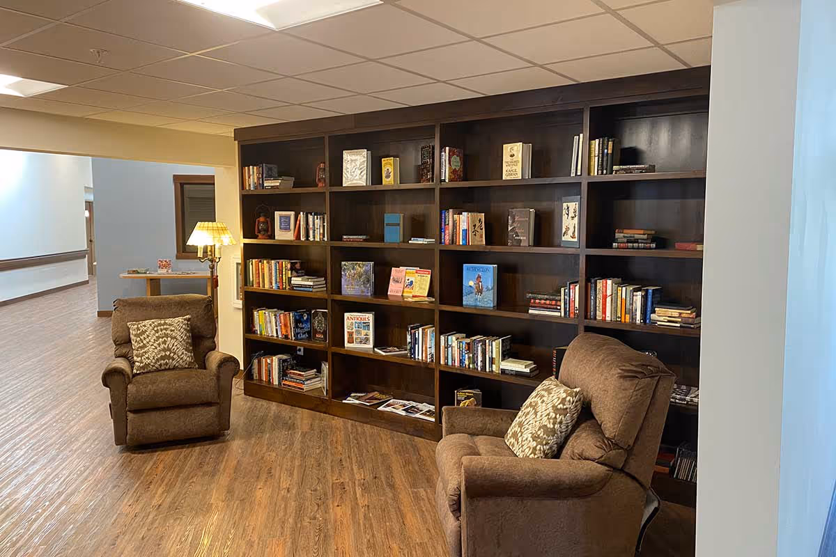 A cozy reading nook in a senior living facility featuring two brown upholstered armchairs with patterned cushions, a tall dark wood bookshelf filled with books and decorative items, a floor lamp with a pleated shade, and wood-look flooring. The area is adjacent to a hallway with handrails and light-colored walls.