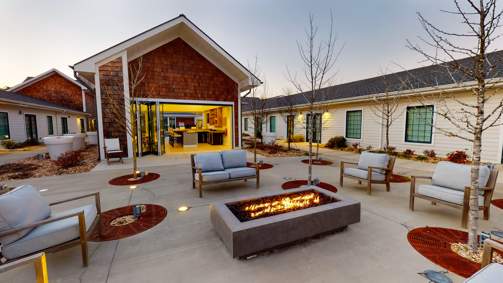 Outdoor courtyard area at Legacy Savannah Quarters featuring a modern rectangular fire pit surrounded by cushioned wooden chairs and small leafless trees planted in circular red mulch beds. The courtyard is paved with concrete and is adjacent to buildings with white siding and large windows. One building has an open area with glass folding doors revealing a kitchen and dining space inside.