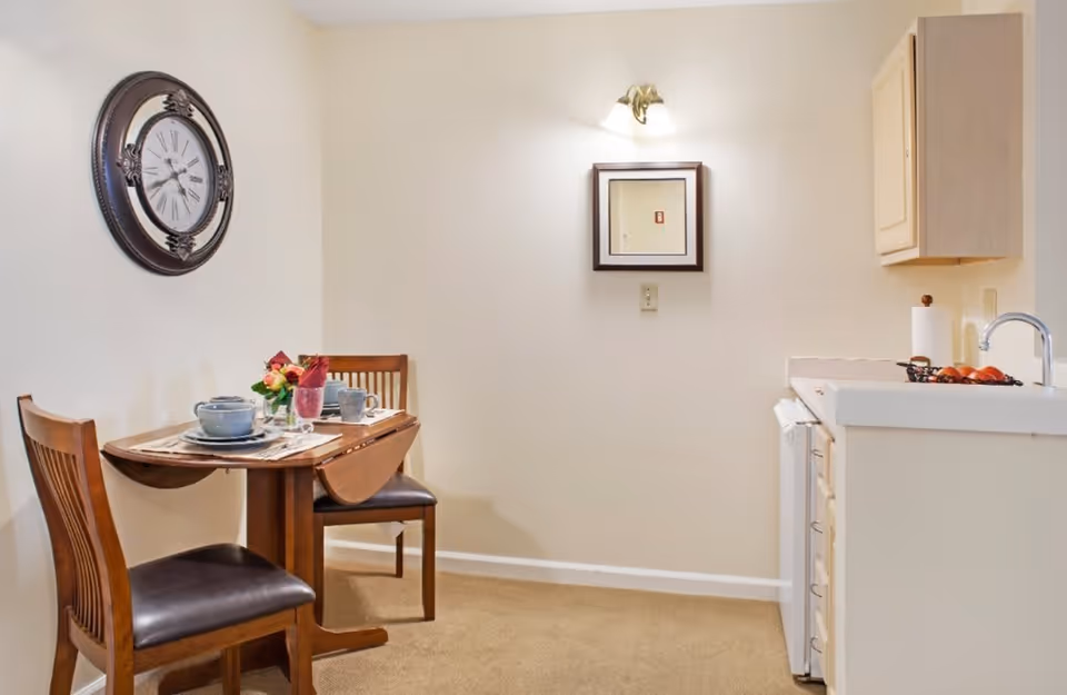 Small dining area with a wooden table set for two with dishes and a flower centerpiece, two wooden chairs with dark cushions, a large wall clock, a wall-mounted light fixture above a small framed mirror, and a kitchenette with a sink, countertop, and cabinets.
