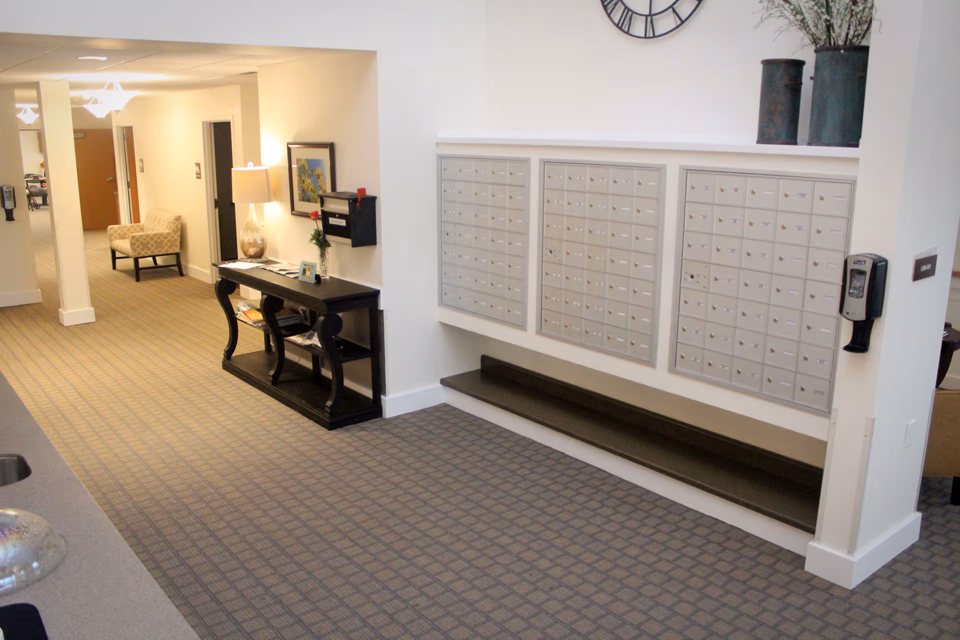 Interior hallway of a senior living facility showing wall-mounted mailboxes, a console table with a lamp and decor, and seating in the background.
