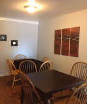 A dining area with two dark wooden tables and several wooden chairs with vertical slats. The walls are light-colored, and there is a three-panel abstract artwork with red and brown tones hanging on one wall. The floor is wooden, and the ceiling light fixture is visible.