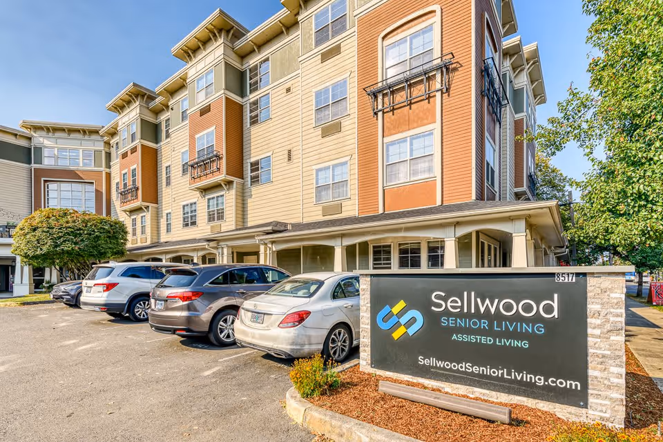 Exterior view of Sellwood Senior Living assisted living facility showing a multi-story building with parked cars in front and a large sign displaying the facility name and website.