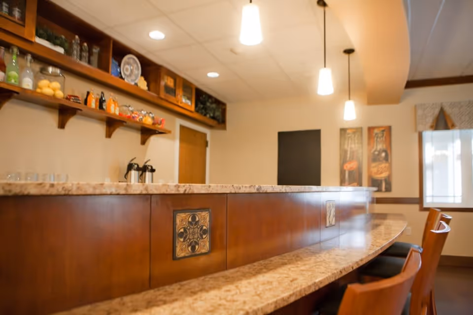 Interior view of a senior living facility's dining or common area featuring a curved marble countertop bar with wooden paneling and decorative tiles. Above the counter are hanging pendant lights, and shelves on the wall hold various bottles and decorative items. There are wooden chairs with black cushions along the counter, and framed artwork and a window with curtains are visible in the background.