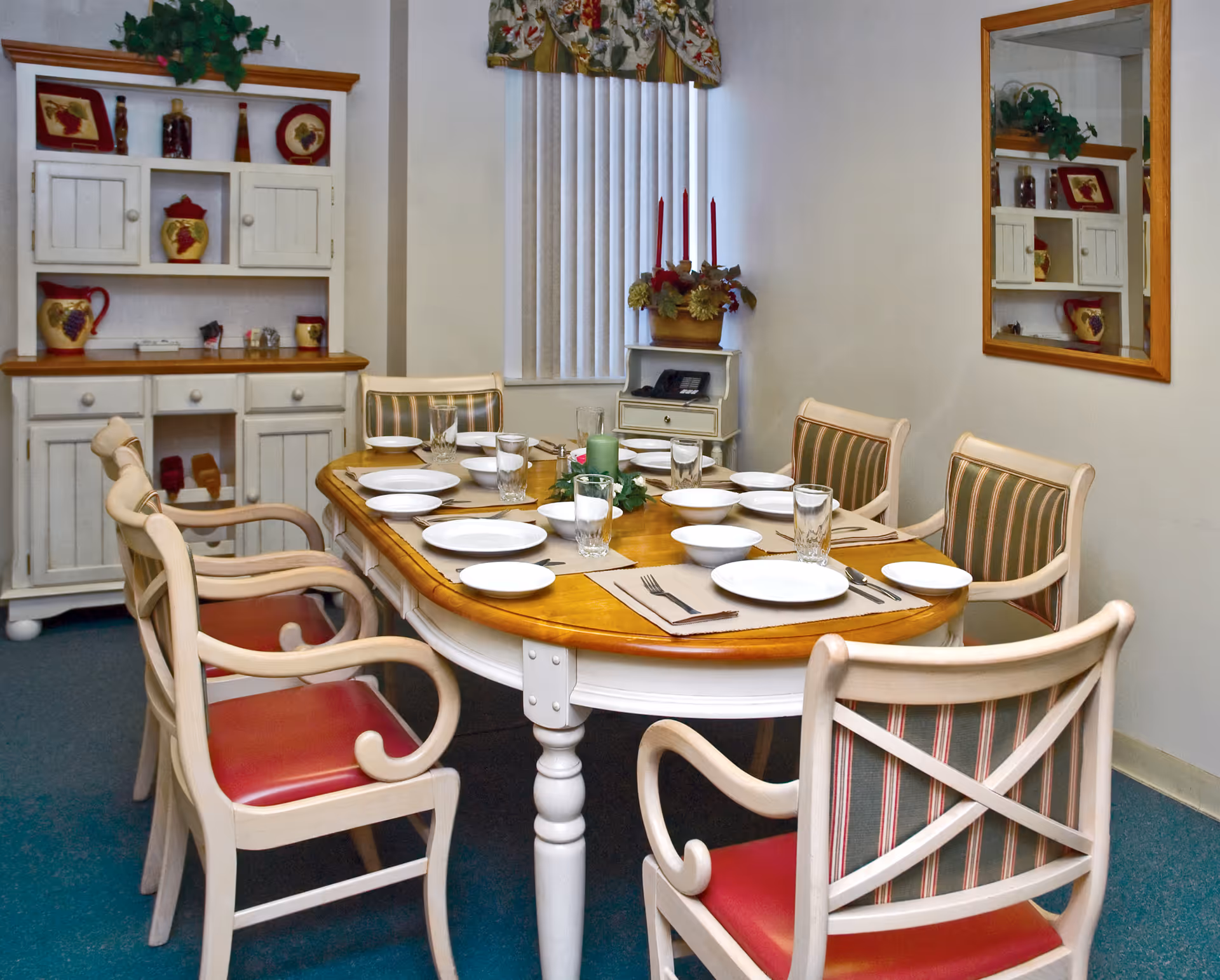 A dining room with a wooden table set for six people, featuring white plates, bowls, glasses, and silverware on beige placemats. The chairs have white frames with red and striped upholstery. In the background, there is a white cabinet with decorative items and a window with vertical blinds and a floral valance. A mirror on the wall reflects part of the room.