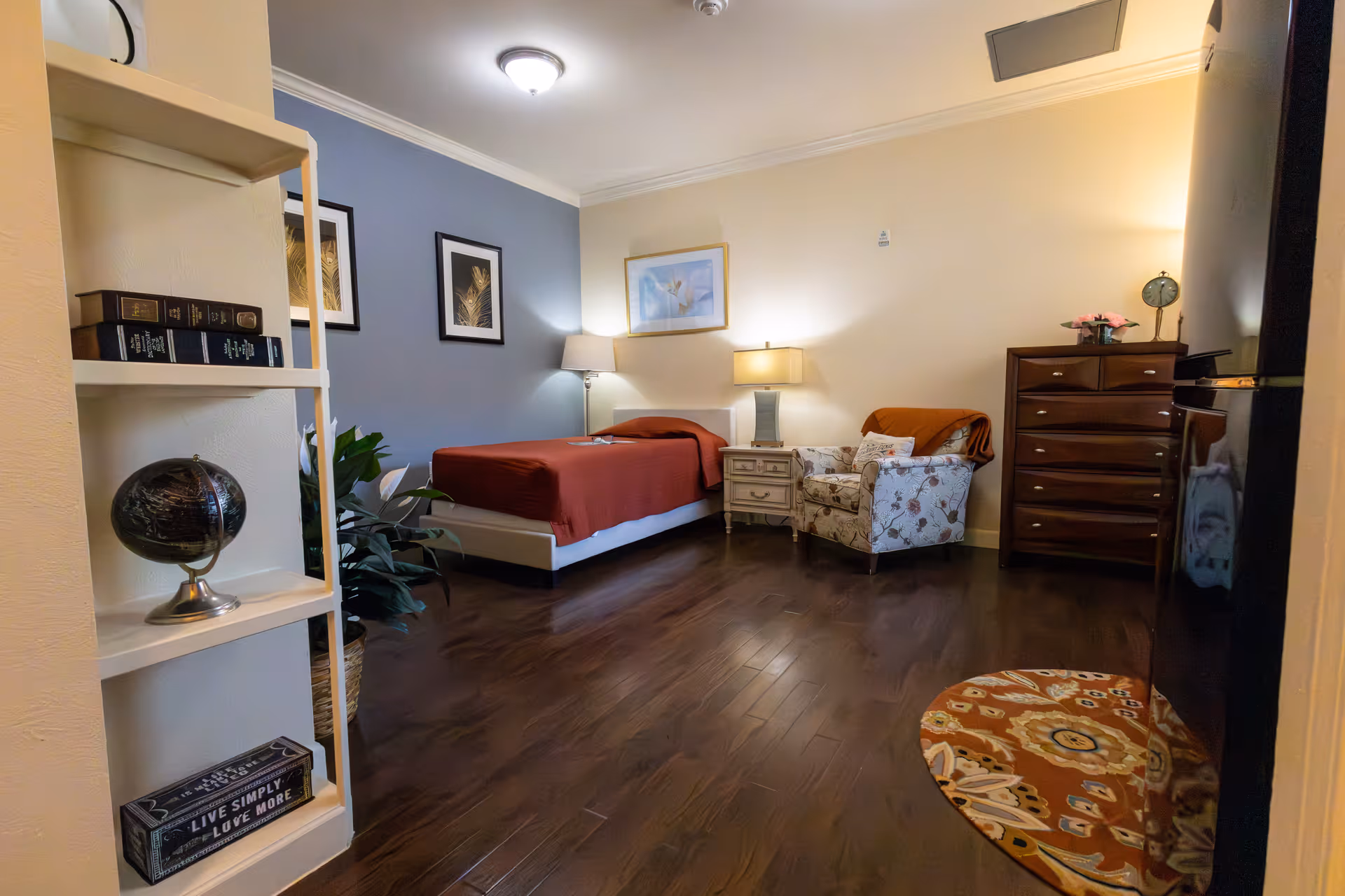 A cozy bedroom in an assisted living facility featuring a single bed with a red bedspread, a nightstand with a lamp, a patterned armchair with an orange throw, a wooden dresser, and a small round rug on the wooden floor. The room also has framed artwork on the walls, a white shelving unit with books and decorative items, and a potted plant.
