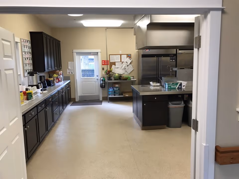 A clean commercial kitchen with dark lower cabinets and countertops along the left, a central island, and stainless steel refrigerators and a door at the back.
