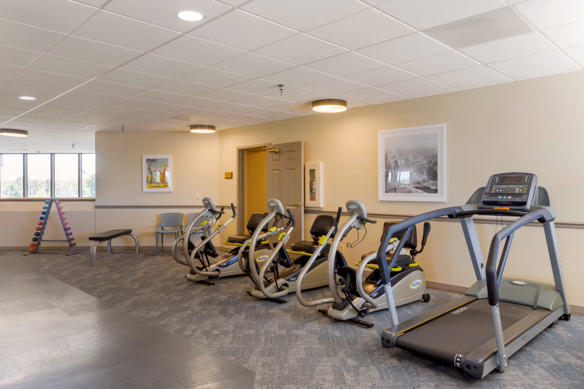 A senior living facility fitness room with exercise equipment including a treadmill, three NuStep recumbent cross trainers, a bench, a chair, and a rack of colorful dumbbells. The room has beige walls, a gray carpeted floor, and large windows letting in natural light. Two framed pictures hang on the walls.