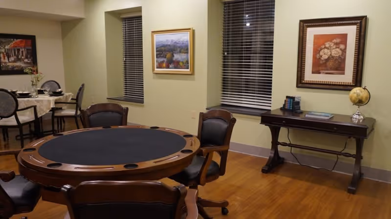 A room with a round poker table surrounded by four leather chairs on wheels. In the background, there is a dining table set with plates and glasses, and two windows with blinds. The walls are decorated with framed paintings, and a wooden console table holds a globe and some books. The floor is wooden.