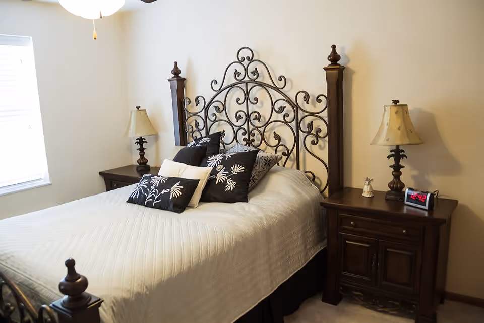 A bedroom with a decorative wrought iron headboard on the bed, which is covered with a white quilt and several black and white patterned pillows. There are two wooden nightstands on either side of the bed, each with a lamp. A digital alarm clock and a small figurine are on the right nightstand. A window with blinds is on the left wall.