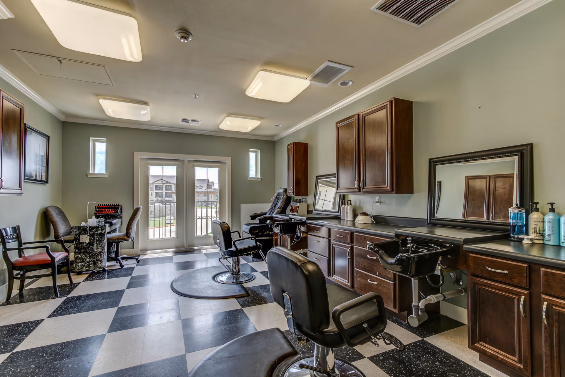 Interior of a salon room with checkered black and white flooring, several salon chairs, a hair washing station, wooden cabinets, mirrors, and a small seating area near double glass doors leading outside.