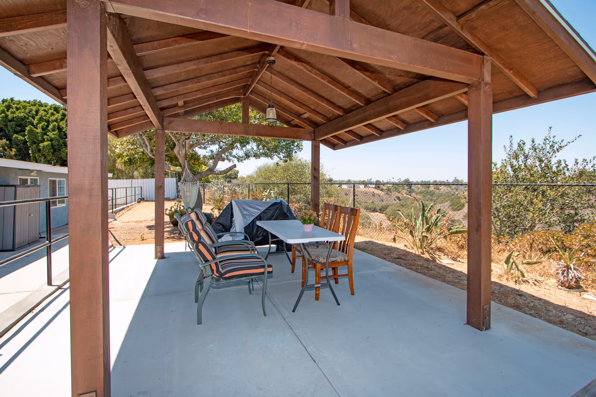 Outdoor covered patio area with a wooden roof structure, featuring a white table surrounded by two cushioned chairs and two wooden chairs. The patio overlooks a natural landscape with trees and shrubs under a clear blue sky.