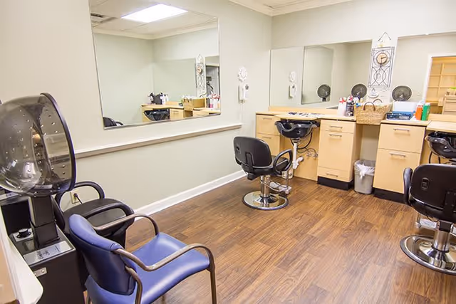 Interior of a hair salon area with three black salon chairs in front of mirrors and wooden cabinets. There are two blue waiting chairs and a hair dryer machine on the left side. The floor is wooden and the walls are light-colored.
