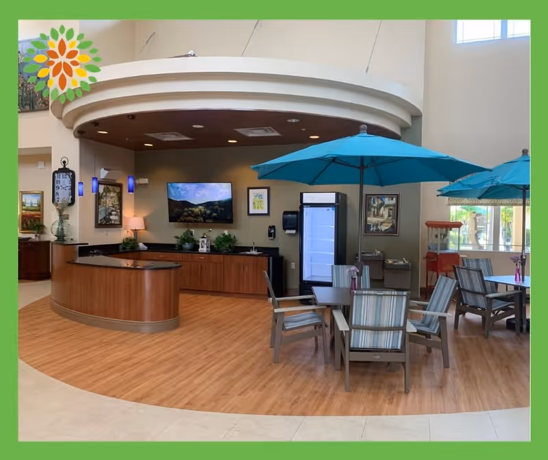 Interior common area with wooden flooring and a curved wooden counter. There are tables with striped cushioned chairs and blue umbrellas indoors. The back wall has a mounted TV, framed pictures, plants, a refrigerator, and a popcorn machine near large windows.