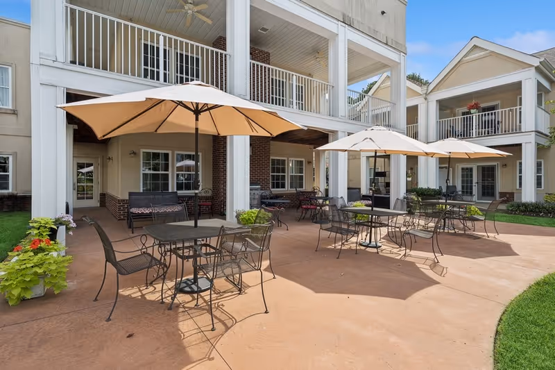 Outdoor patio area at Park Place of West Knoxville with several metal tables and chairs under large beige umbrellas. The patio is adjacent to a building with balconies and white columns. There are potted plants and green grass surrounding the patio.