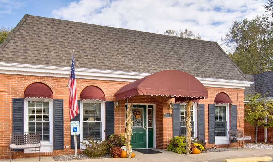 Brick senior living facility entrance with green door, maroon awnings, flag, pumpkins, and benches.