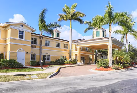 Front entrance of a yellow two-story senior living building with palm trees and a covered porte-cochere.