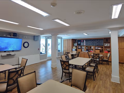 A well-lit common area with multiple tables and chairs arranged for group activities. A large TV is mounted on the wall to the left, with a clock nearby. The back wall features shelves and a large bingo board. The floor is wooden, and there are windows and doors letting in natural light.