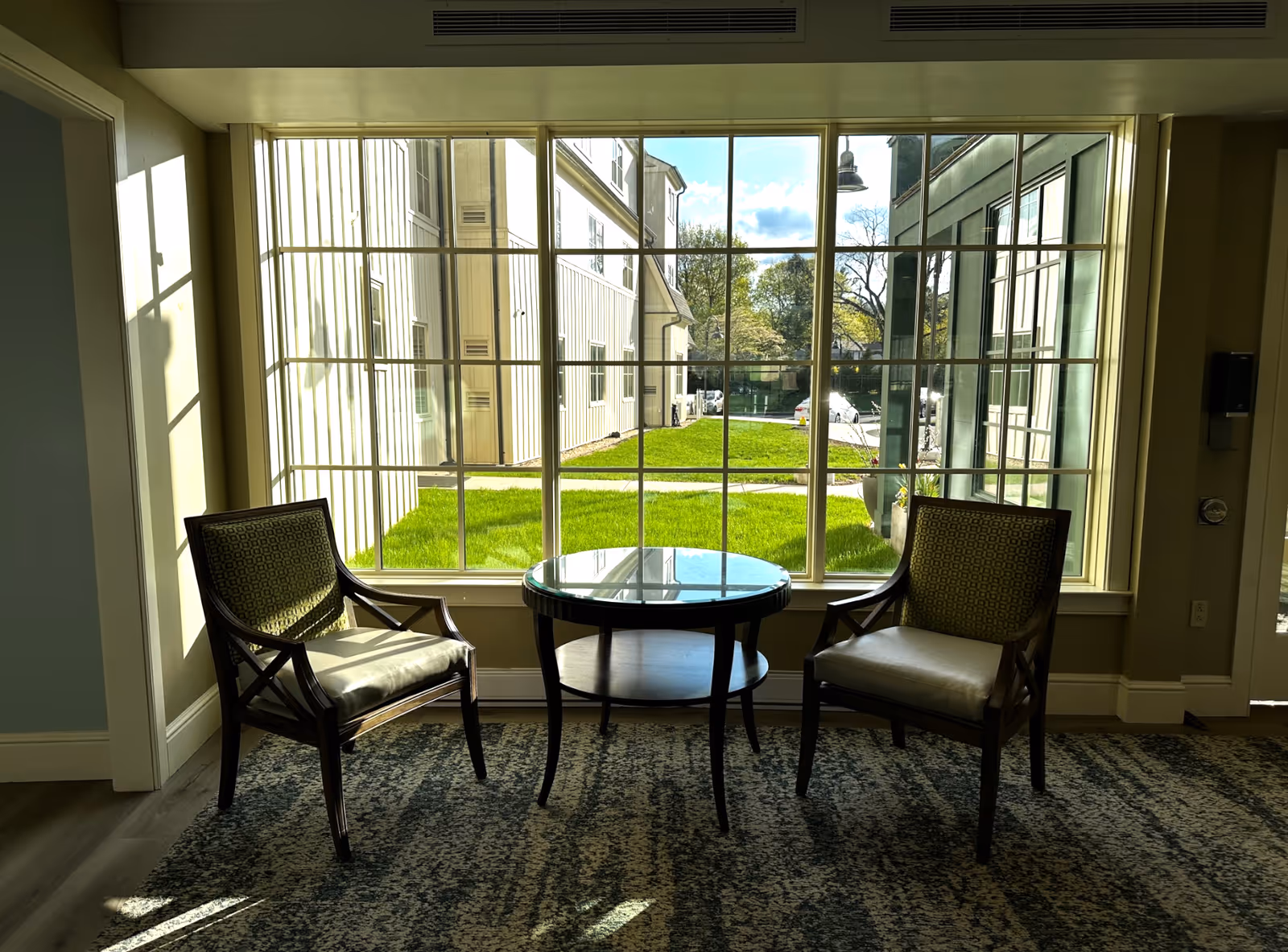 Two wooden armchairs with patterned cushions are positioned on either side of a small round glass-top table in front of a large window. Outside the window, there is a view of green grass, a sidewalk, and parts of adjacent buildings under a blue sky with some clouds. The interior has a carpeted floor and neutral-colored walls.
