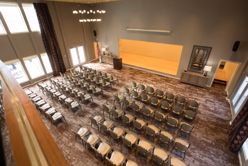 A large room set up for a presentation or event with rows of chairs facing a small stage area. The room has tall windows with curtains, a chandelier, and neutral-colored walls and carpet. There is an American flag and a podium near the stage.