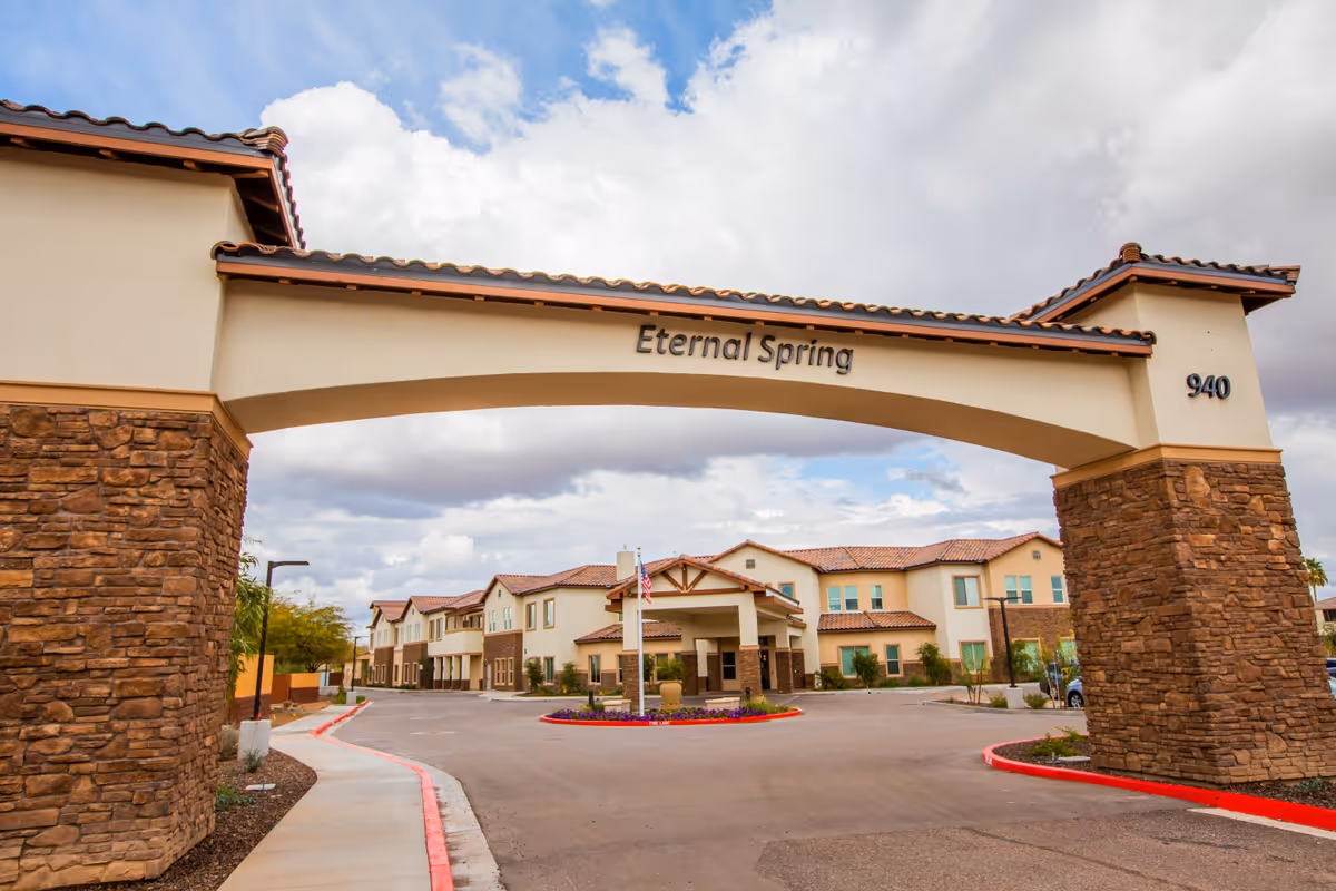 Entrance archway with stone pillars and a tiled roof at the senior living facility named Eternal Spring. The archway has the name 'Eternal Spring' and the number '940' displayed. Behind the archway, there is a two-story building with a similar tiled roof and beige exterior walls. The sky is partly cloudy.