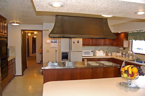 Interior view of a kitchen with wooden cabinets, a large range hood above a stove, a refrigerator, microwave, toaster, and various kitchen appliances on the countertops. A bowl of fruit is placed on a counter in the foreground, and a window with a valance is visible on the right side.