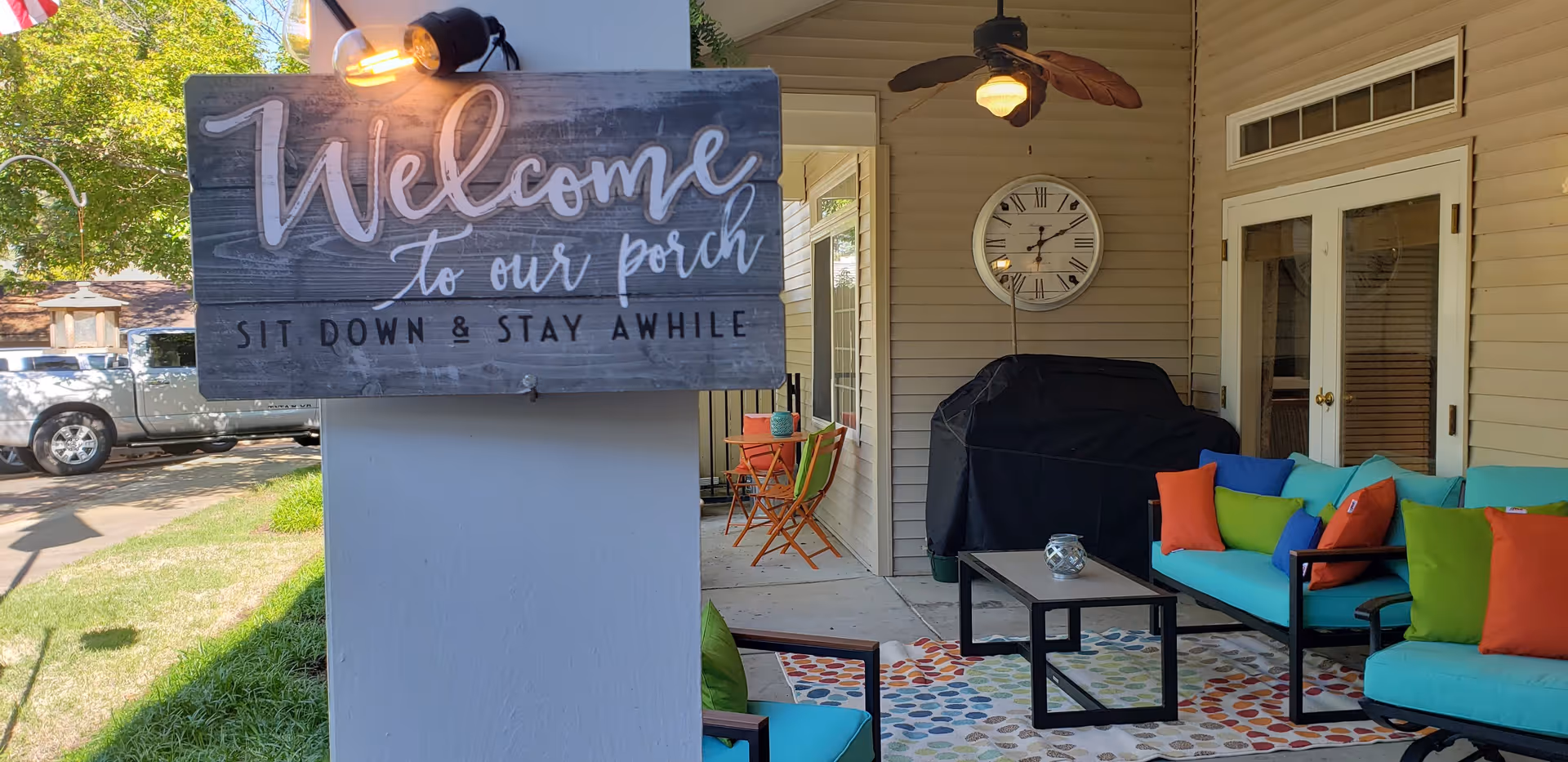 Covered porch with a 'Welcome to our porch' sign, colorful seating, a coffee table, a grill, and a wall clock.