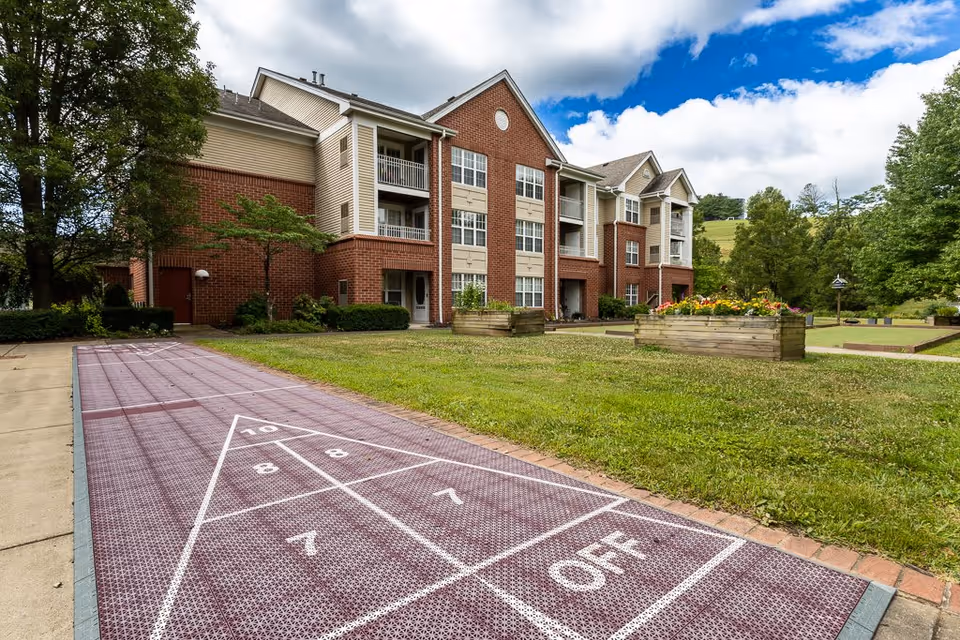 Outdoor area of a senior living facility featuring a shuffleboard court in the foreground, a well-maintained lawn with flower beds, and a three-story brick and siding building with balconies in the background under a partly cloudy sky.