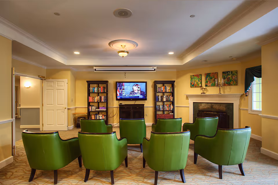 A cozy common room with eight green armchairs arranged in two rows facing a wall-mounted television. The room features two bookshelves filled with books on either side of the TV, a fireplace with decorative artwork above it, and soft yellow walls with white trim. There is a window with a blue valance on the right side and an open door leading to another room on the left.