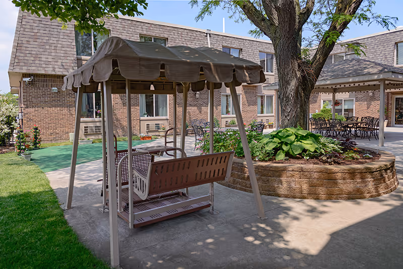 Outdoor patio area at Rivergate Terrace featuring a covered swing bench, a large tree surrounded by a raised brick planter with greenery, and several tables and chairs under a gazebo. The building's brick exterior and windows are visible in the background.