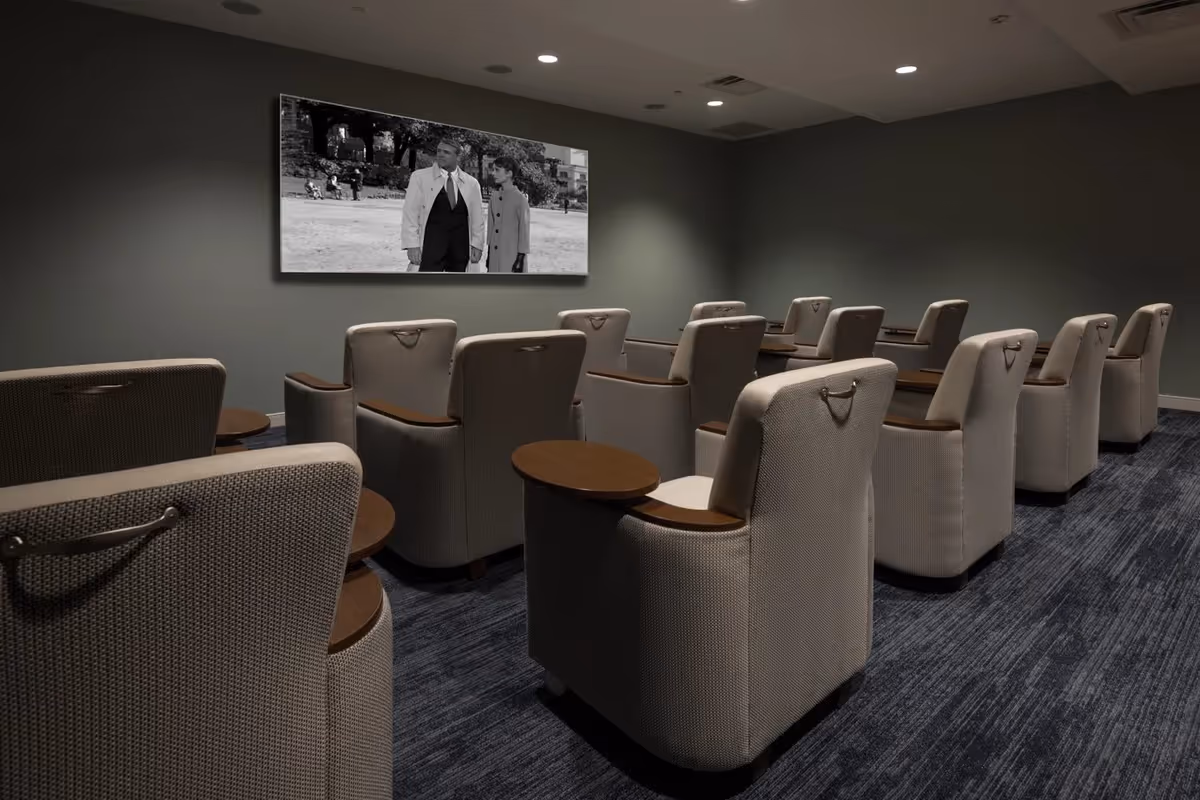 Small private theater room with rows of upholstered recliner chairs facing a wall-mounted screen showing a black-and-white movie.