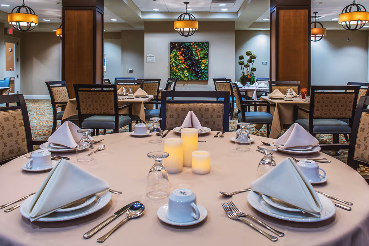 A neatly set dining table in a senior living facility dining room with white plates, folded napkins, cups, glasses, and silverware arranged around the table. Three decorative candles are placed in the center. The room has multiple tables and chairs, warm lighting fixtures, and a colorful plant wall decoration in the background.