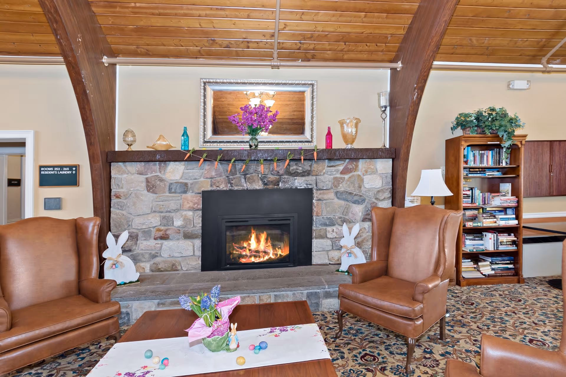 Cozy living room area with a stone fireplace featuring a burning fire. The mantel is decorated with a mirror, purple flowers, colorful bottles, and a garland of carrots. Two brown leather armchairs flank the fireplace, and a wooden coffee table with a floral centerpiece and Easter decorations is in the foreground. A bookshelf filled with books and a lamp are visible to the right, and a sign indicating room numbers and laundry is on the left wall.