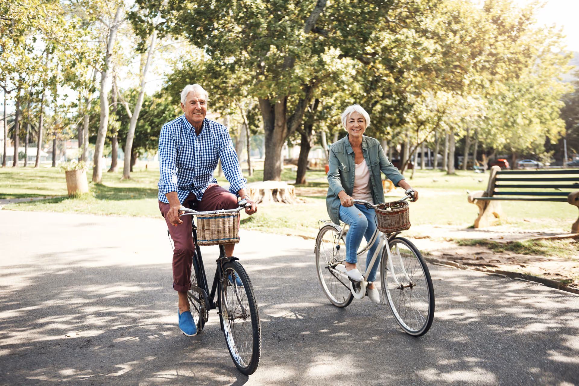 An elderly man and woman riding bicycles on a paved path in a park with trees and benches around them, enjoying a sunny day.