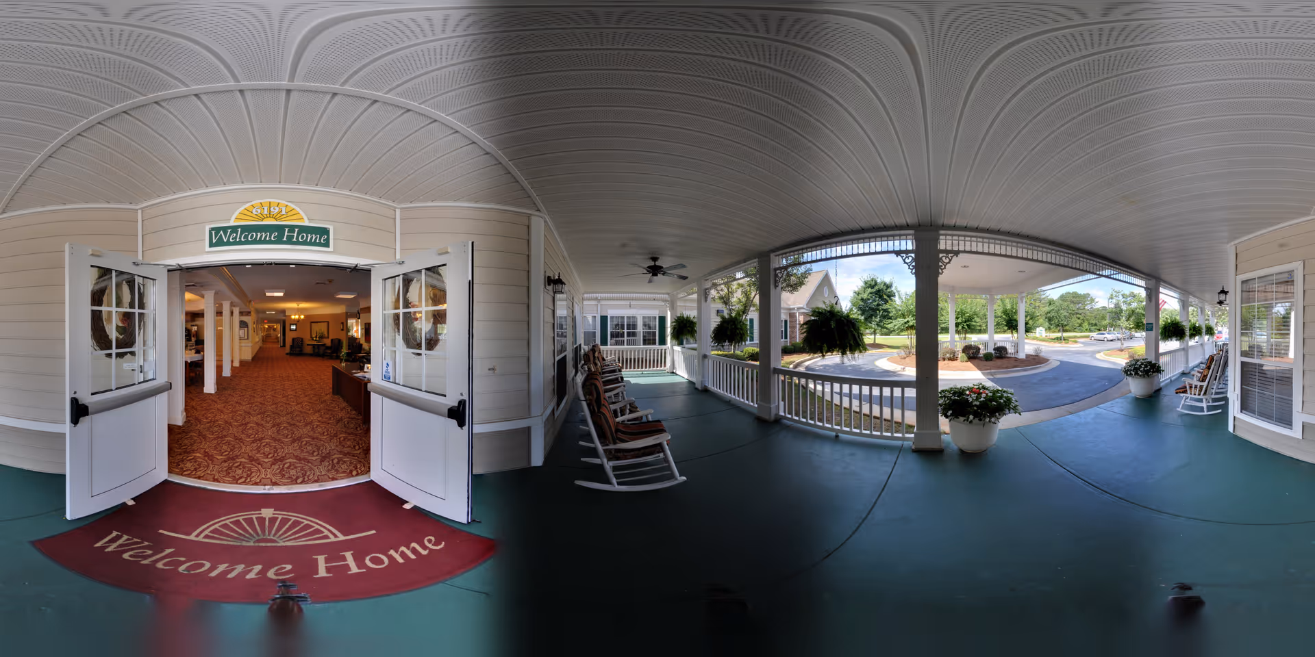 Covered entrance area of a senior living facility with white rocking chairs and potted plants on a green floor. Double doors open inward to a warmly lit interior with carpeted flooring and a sign above the doors that reads 'Welcome Home'. Outside, there is a circular driveway and landscaped greenery under a partly cloudy sky.