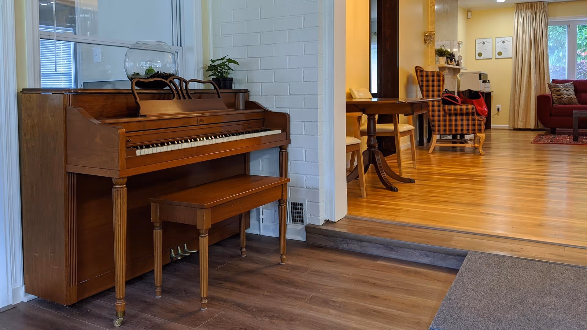 Upright wooden piano and bench by an open doorway into a wood‑floored living area with chairs and a sofa.