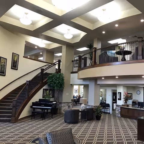 Spacious senior living facility lobby with a curved staircase, a black piano, patterned carpet, seating areas with chairs and tables, and a second-floor balcony overlooking the area. The space is well-lit with ceiling lights and decorated with plants and framed artwork.