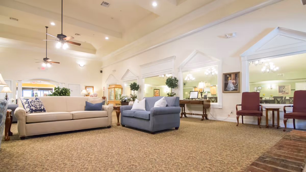 A spacious and well-lit living room area with a beige sofa and a blue loveseat, both adorned with decorative pillows. The room features carpeted flooring, ceiling fans with lights, and large windows with white trim. There are also two red chairs with a small table between them, a wooden side table with a lamp, and some greenery including potted plants. The walls are light-colored, and there are framed pictures and mirrors adding to the decor.