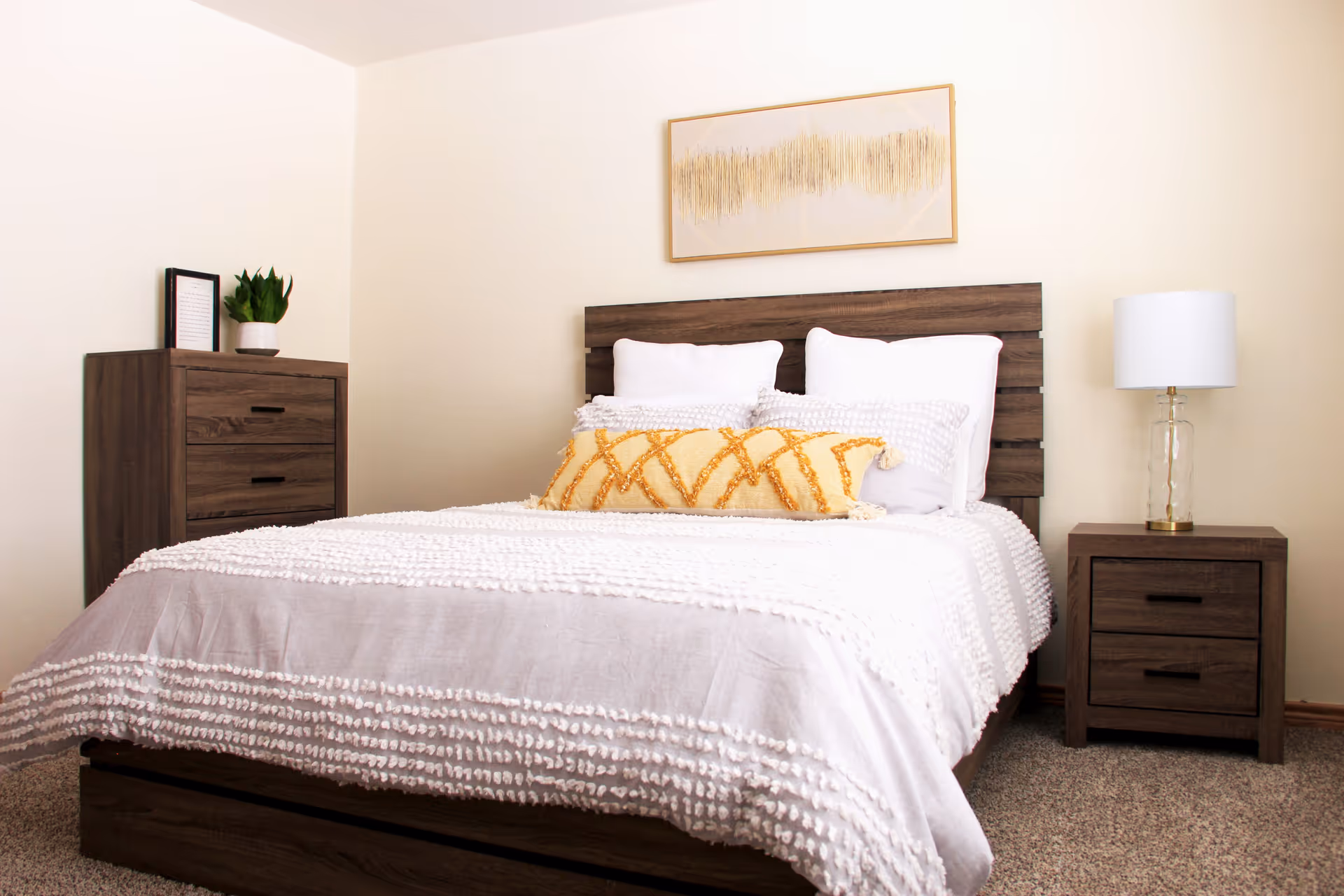 A neatly made bed with white textured bedding and a yellow decorative pillow in a bedroom. The bed has a wooden headboard and is flanked by a wooden nightstand with a glass lamp on the right and a wooden dresser with a small potted plant and framed picture on the left. A framed abstract artwork hangs on the wall above the bed.