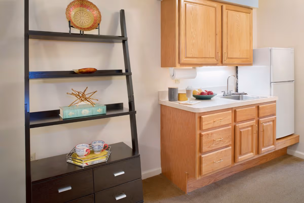 Small kitchenette with oak cabinets, a sink and refrigerator next to a black ladder-style shelf displaying decorative items.